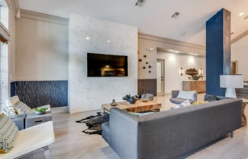 Modern living room in The Highlands Reno features a gray sectional sofa, wooden coffee table, wall-mounted TV on a marble accent wall, and contemporary decor. Natural light pours in through large windows on the left.