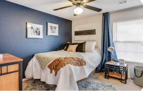 A cozy bedroom in The Highlands Reno features a bed against a dark blue accent wall, adorned with framed photos, a side table with a lamp, and a ceiling fan overhead. Blue curtains on the window let in natural light.