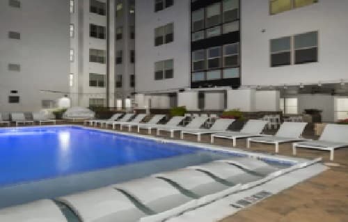 Blurry image of a modern outdoor swimming pool surrounded by white lounge chairs, set in the courtyard of The Hudson apartments in College Station.