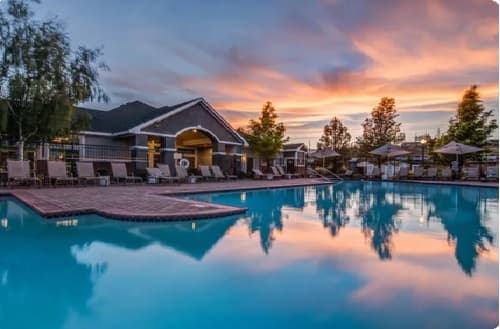A serene outdoor swimming pool at The Highlands Reno reflects a colorful sunset sky, with lounge chairs and umbrellas lining the poolside near a building surrounded by trees.