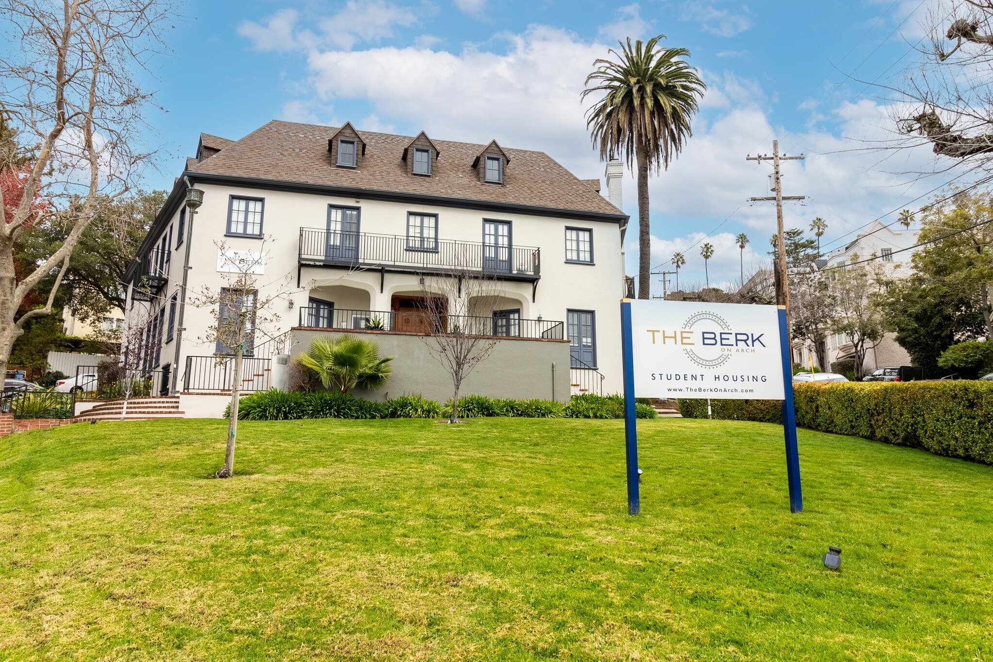 A large, three-story house with dormer windows and a sign in front reading The Berk Student Housing sits on a green lawn, surrounded by palm trees, under a partly cloudy sky.