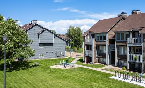 A sunny outdoor scene at The Junction Apartments Fort Collins shows two buildings with brown roofs, grassy lawns, a picnic table, sidewalks, trees, and a bike rack under a blue sky with scattered clouds.
