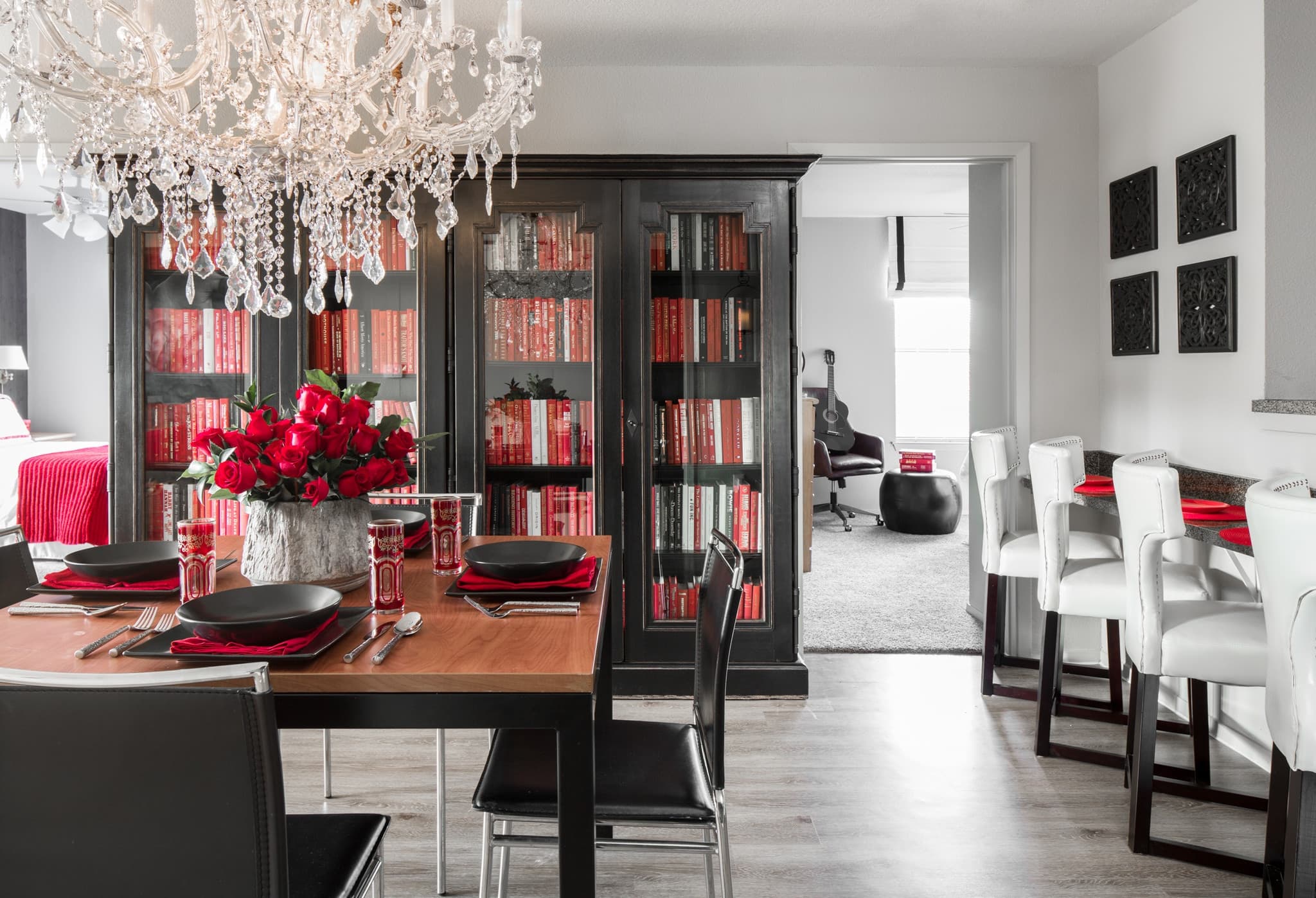 A modern dining room with a wooden table set for four, black and red dishes, a crystal chandelier, black bookshelves filled with red books, and a flower centerpiece. A kitchen and sitting room are visible in the background.