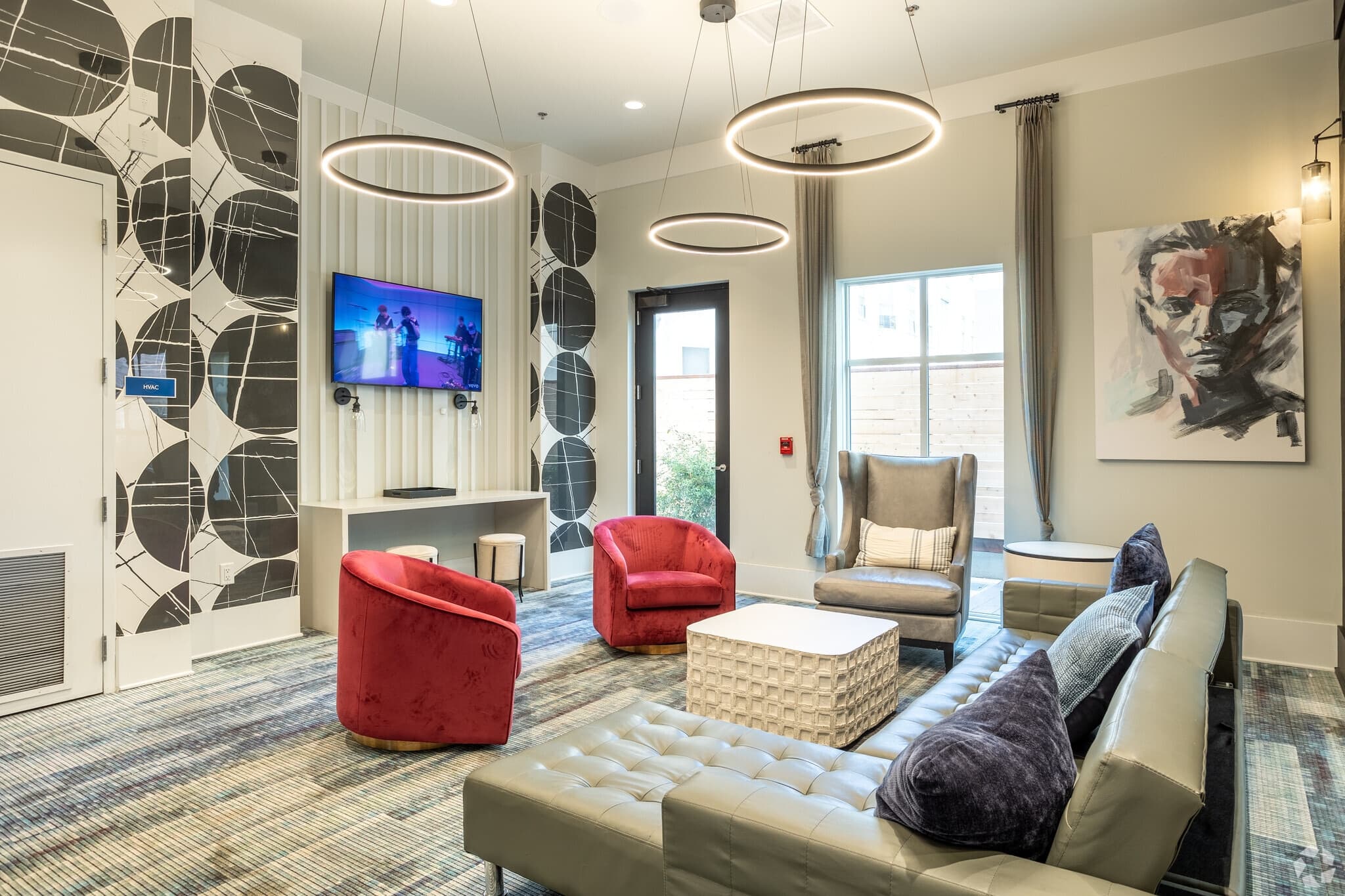 Modern lounge at The Standard at Berkeley with a gray sectional sofa, two red chairs, an armchair, a TV on the wall, abstract wall art, circular pendant lights, large windows with curtains, patterned carpet, and stylish wall accents.