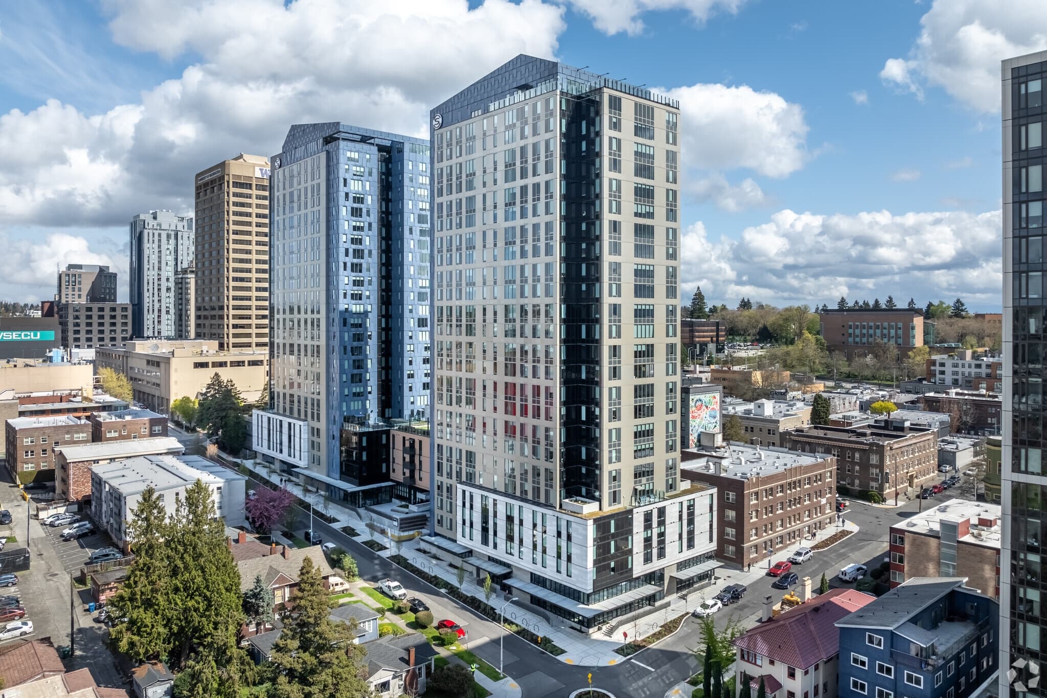 Aerial view of The Standard at Seattle, two modern high-rise apartment buildings in a city neighborhood, surrounded by smaller buildings, trees, and streets under a partly cloudy sky.