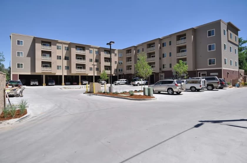 An uncommon four-story beige apartment building in Fort Collins with balconies, a parking lot out front, several parked cars, some landscaping with small trees and shrubs, and a clear blue sky overhead.