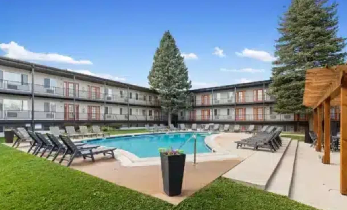 A circular outdoor swimming pool at The Lodge Boulder is surrounded by lounge chairs and a three-story apartment building with balconies. A large evergreen tree, lush grass, and a wooden pergola sit to the right under a clear blue sky.