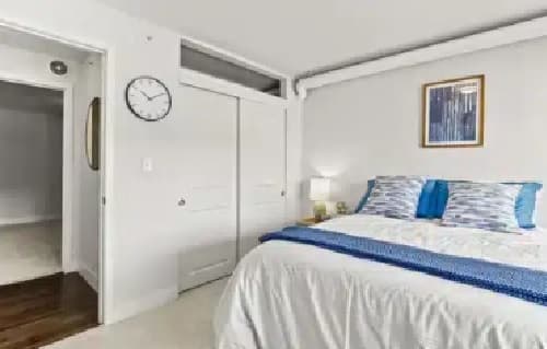 A modern bedroom at The Lodge Boulder features a neatly made bed with blue and white bedding, a nightstand with a lamp, a wall clock, a closet with double doors, and a framed picture on the white wall.