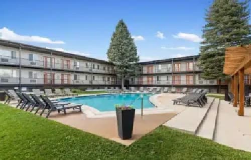A courtyard at The Lodge Boulder features a swimming pool surrounded by lounge chairs, grass, and a three-story apartment building. A tall pine tree and a wooden pergola add charm to the inviting outdoor space.
