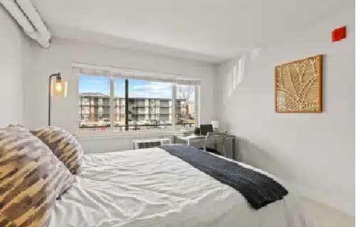 Bright bedroom at The Lodge Boulder with a large window, double bed with patterned pillows and a dark throw, small desk and chair, wall art, and views of apartment buildings. Room features white walls and minimal decor.
