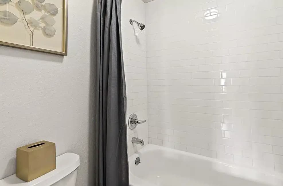 A white bathtub with chrome fixtures is set against a white subway tile wall at The Lodge Boulder. A dark gray shower curtain is partially closed, and a framed leaf print hangs above a toilet with a gold tissue box on top.