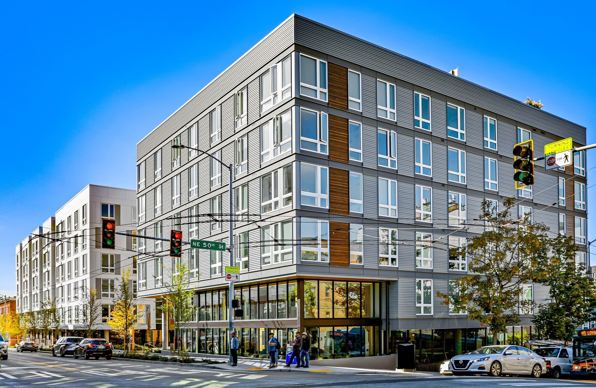 A modern, multi-story apartment building with large windows rises in the heart of Theory U District, featuring street-level businesses at a bustling city intersection with traffic lights, crosswalks, and pedestrians enjoying a sunny day.
