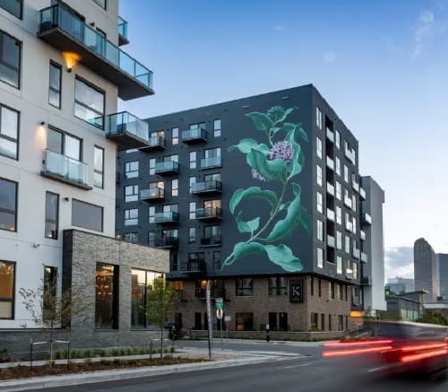 A modern apartment building with balconies, featuring a striking mural of green leaves and a purple flower by Kendrick on a dark wall. The city skyline rises in the background under a blue sky, while cars are parked on the street below.