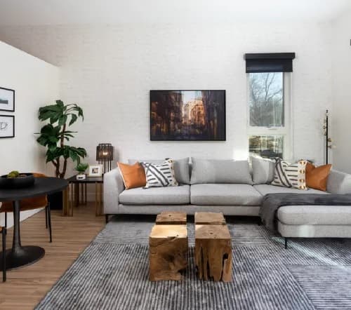 Modern living room with a light gray sectional sofa, striped and patterned pillows, two rustic wooden coffee tables, large Kendrick wall art, a round dining table, indoor plant, and natural light from a window.