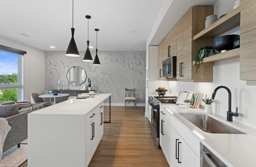 Modern kitchen with white cabinets, wood accents, and a large island. Black Kendrick pendant lights hang above the island. In the background, there’s a dining area with a round table, chairs, and a textured accent wall.