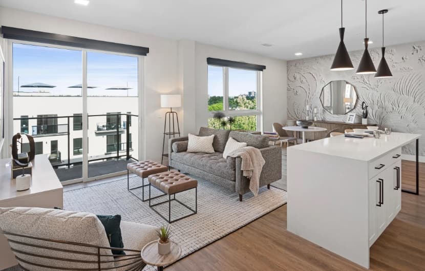 Modern open-concept living room and kitchen with neutral decor, large windows, balcony view, cozy seating, a light rug, and a Kendrick-inspired kitchen island with pendant lights. Dining area and decorative wall are visible in the background.