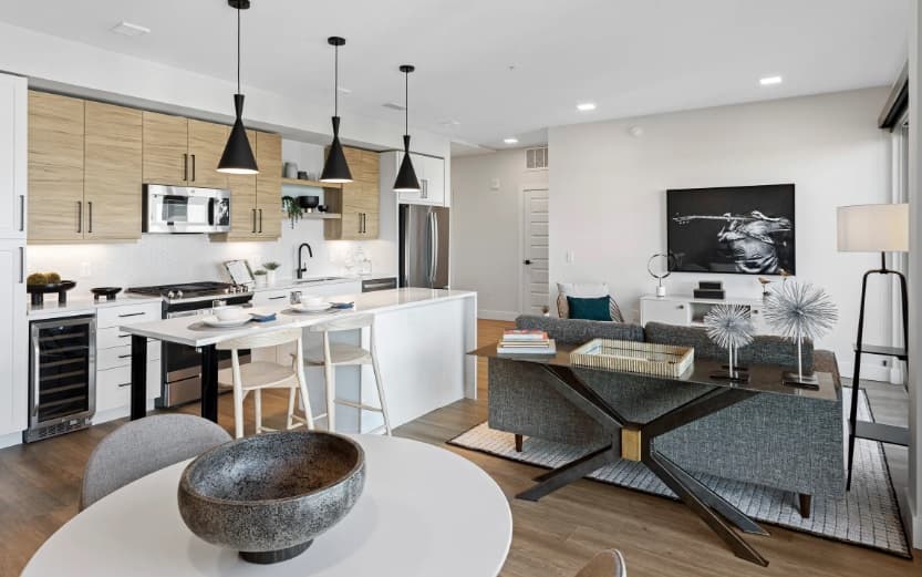 Modern open-plan kitchen and living area with light wood cabinets, white countertops, and black pendant lights. A dining table sits in the foreground, while a gray Kendrick sofa faces a TV and artwork on the wall.