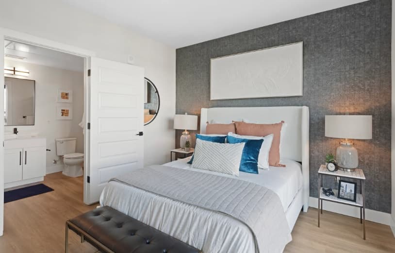 Modern bedroom with a gray accent wall, white upholstered bed, blue and beige pillows, and two bedside tables with lamps—channeling a subtle Kendrick vibe—plus an open door leading to a bright bathroom.