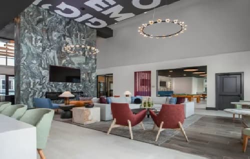 Modern lounge area in The Landing College Station features colorful chairs, sofas, two circular chandeliers, a marble accent wall with a TV, and tables; pool tables and extra seating complete this bright, contemporary student housing space.