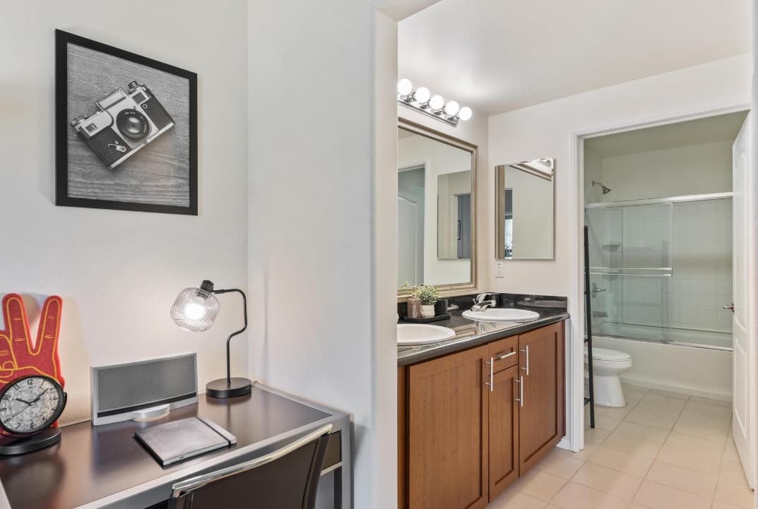 A modern bathroom with double sinks and a glass shower door is adjacent to a small desk setup with a lamp, clock, and framed camera photo on the wall.