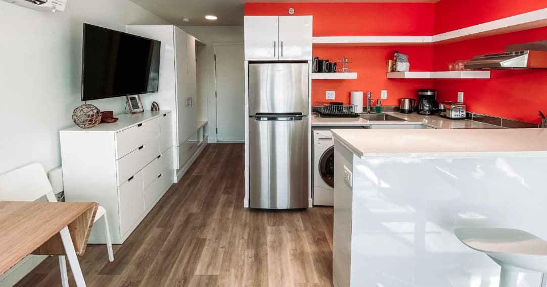Modern kitchen at The Mod at Riverwalk Reno with white cabinets, stainless steel fridge, and an orange accent wall. Open shelves display kitchen items, a mounted TV sits to the left, and light wood flooring leads to a small dining table.