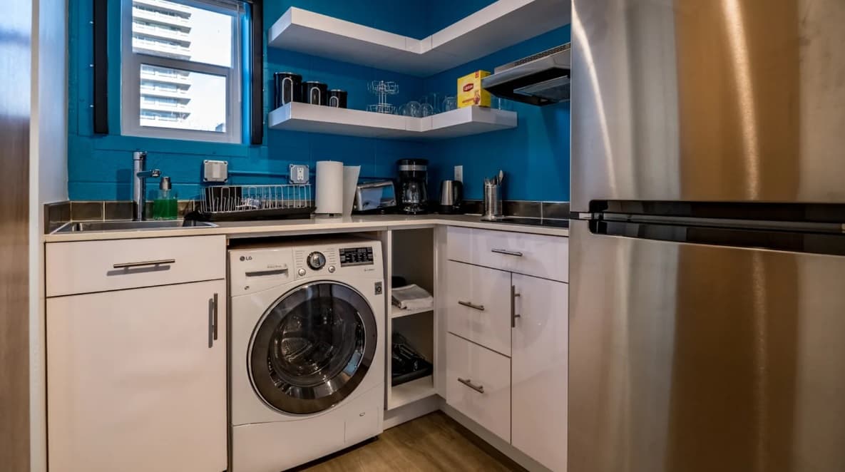 A modern kitchen at The Mod at Riverwalk Reno features blue walls, white cabinets, a washing machine, stainless steel refrigerator, countertop appliances, and open shelves for dishes. A window fills the space with natural light.