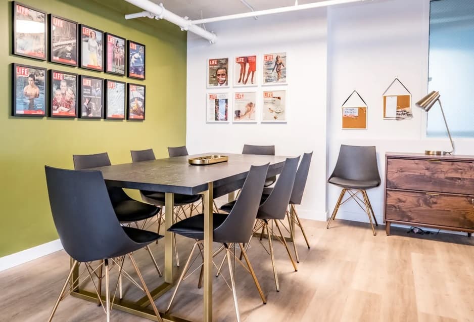 Modern meeting room at The Mod at Riverwalk Reno with a black table, eight black chairs with gold legs, framed magazine covers on green and white walls, a wooden cabinet, corkboards, and light wood flooring.