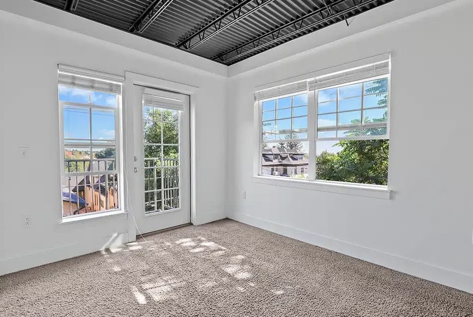 Bright, empty room with white walls, large windows, and a glass door leading to a small balcony at The Mansion Boulder. Exposed black ceiling beams and sunlight streaming onto the light brown carpeted floor create an inviting space.