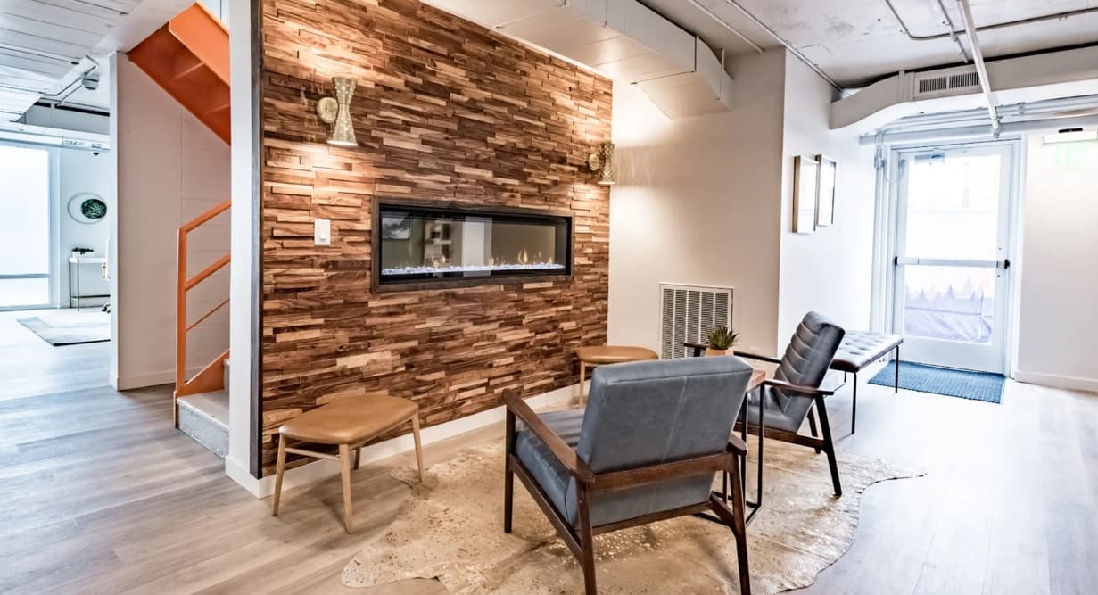 Modern lounge area at The Mod at Riverwalk Reno with wood-paneled fireplace, two gray armchairs, a stool, and a small table on a cowhide rug. An orange staircase and light wood flooring lead to an open, bright hallway.