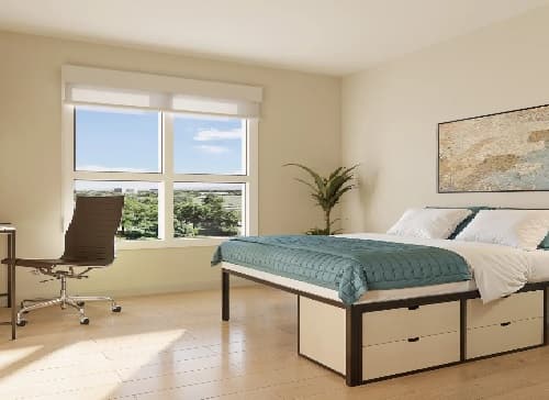 A bright, minimalist bedroom in Mark Fort Collins features a large window, a neatly made bed with teal bedding, a wooden desk and chair, a potted plant, and storage drawers under the bed. Sunlight fills the room.