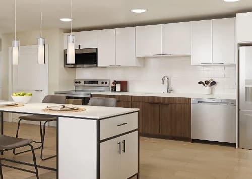 Modern kitchen with white upper cabinets, wood lower cabinets, stainless steel appliances, a kitchen island with two chairs, pendant lights, and a bowl of green apples—an inviting space in the heart of Mark Fort Collins.