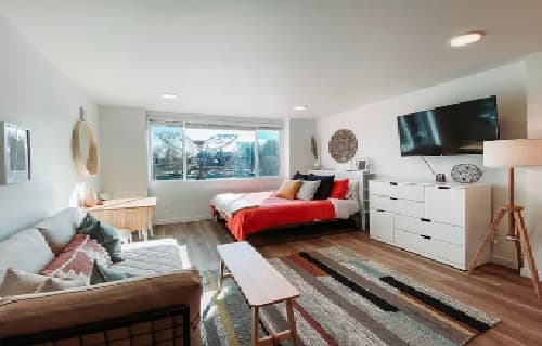 Modern, brightly lit studio apartment at The Mod at Riverwalk Reno with a bed, sofa, TV on the wall, dresser, floor lamp, large window, and colorful decor accents including cushions and a striped rug.