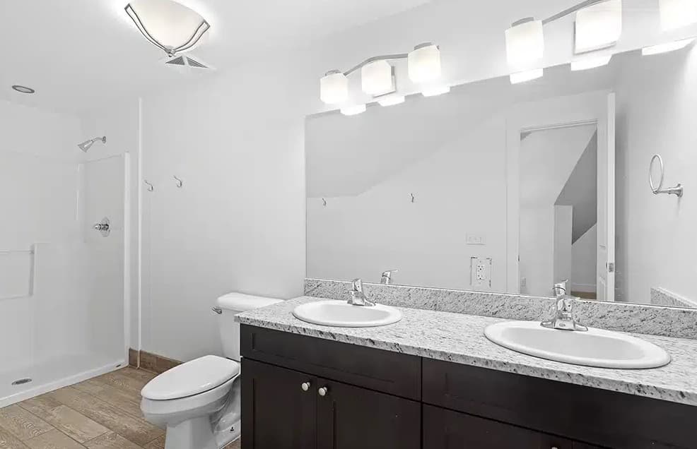 Modern bathroom at The Mansion Boulder with a large mirror, double sink vanity featuring a granite countertop, white toilet, walk-in shower, and wood-look tile flooring. Bright lighting fixtures illuminate the sinks.
