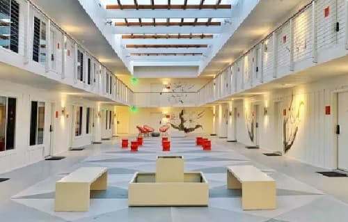 A bright, modern indoor space at The Mod at Riverwalk Reno features white walls, benches, and red chairs arranged in the center. Natural light streams through a skylight onto painted plants, creating a welcoming communal lounge area.
