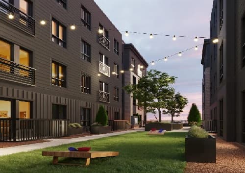 A modern apartment courtyard at dusk at Mark Fort Collins, featuring string lights overhead, green grass, bean bag toss games, potted plants, and seating areas between two dark-colored student apartment buildings in Colorado.