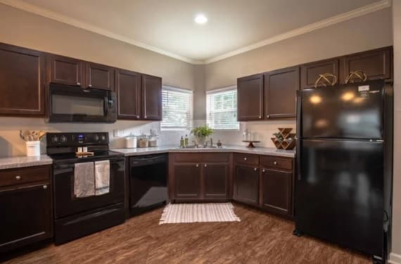 Modern kitchen at The Outpost Fort Collins features dark wood cabinets, black appliances, a corner sink under two windows, stylish countertop decor, a small rug on brown wood floors, and neutral walls with crown molding.