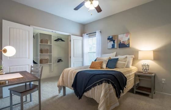 A cozy bedroom at The Outpost Fort Collins features a neatly made bed with a navy throw, pillows, bedside tables with lamps, an open closet with baskets and hangers, a desk with a lamp, and a window letting in natural light.