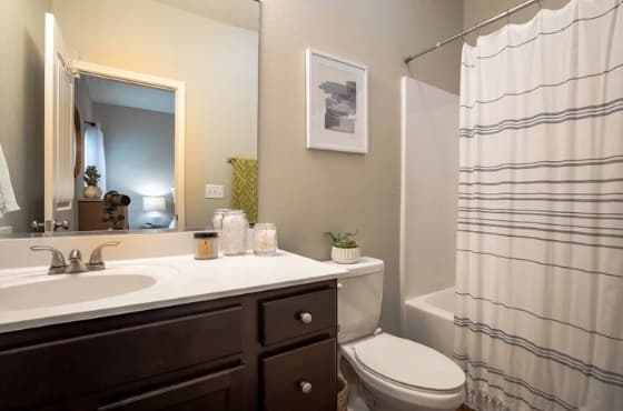 A clean bathroom at The Outpost Fort Collins features a dark wood vanity, white countertop, sink, toilet, and a bathtub with a white and gray striped shower curtain. A framed picture hangs above the toilet, and a mirror reflects a bedroom.