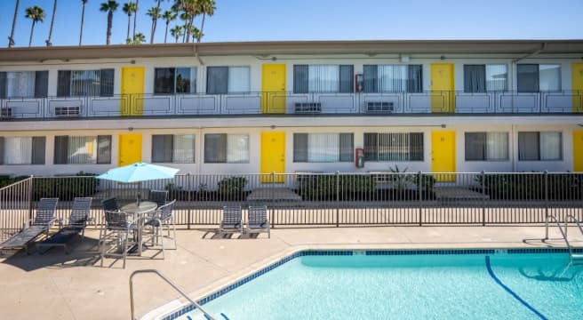 A small outdoor swimming pool is surrounded by a metal fence, with lounge chairs and a table with a blue umbrella nearby. In the background is a two-story motel with yellow doors, palm trees, and an SDSU Toltec vibe.