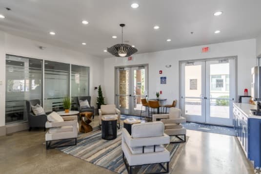 Modern lounge area at The Republic Reno with beige armchairs, small round tables, a blue-striped rug, and a chandelier. Large windows let in natural light; a glass door and small dining set appear in the background.