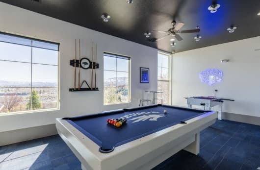 A game room at The Republic Reno with a white pool table featuring the Nevada Wolf Pack logo, billiard balls arranged on the table, a foosball table, wall-mounted pool cues, Nevada decor, and large windows showcasing views of trees and mountains.