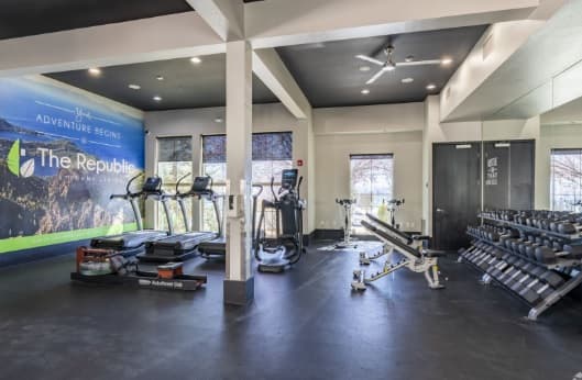 A modern gym at The Republic Reno with treadmills, elliptical machines, a weight bench, and a rack of dumbbells. Large windows flood the space with natural light, and a wall mural reads Adventure Begins - The Republic.
