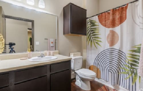 Modern bathroom at The Republic Reno with dark wood cabinets, a white sink, and a shower with a colorful curtain featuring abstract shapes and leaf patterns. Towels and toilet sit beside the sink, beneath a large mirror above.