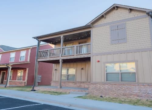 A two-story house with tan and brown siding, a covered front porch, and a second-floor wooden balcony—perfect for student housing. The house has large windows and sits next to a red-painted home on a residential street.