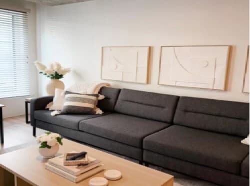 A modern living room with a dark gray sofa, neutral throw pillows, and a blanket. Above the sofa are three minimalist framed art pieces. A wooden coffee table with a vase and books sits in front, and light streams through window blinds.