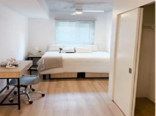 A bright, minimalist bedroom with a large bed under a window with blinds, a wooden desk and chair on the left, and an open closet with sliding doors on the right. The floor is light wood.