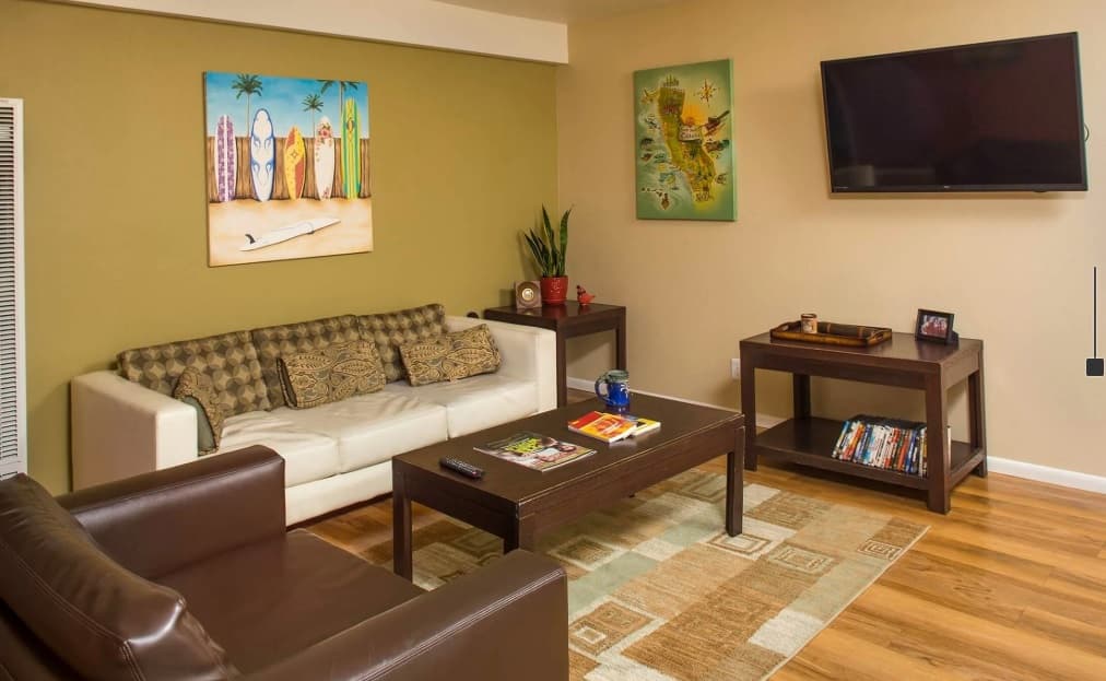 A cozy living room in Tropicana Gardens with a white sofa, brown armchair, two wooden coffee tables, wall art, a plant, and a flat-screen TV. Magazines and books are neatly placed on the tables and shelf.