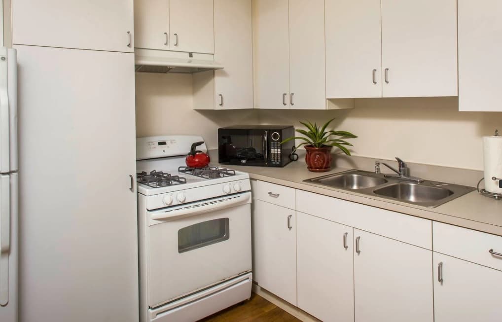 A small kitchen in Tropicana Gardens features white cabinets, a white gas stove with a red kettle, a refrigerator, microwave, double sink, potted plant, and paper towel holder on the counter.
