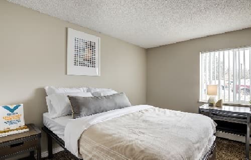 A neatly made bed with white and beige bedding sits by a window with vertical blinds at The Social at Stadium Walk Fort Collins. Above the bed hangs a painting, while a small desk with a lamp and books sits to the right. The room features neutral walls and carpet.