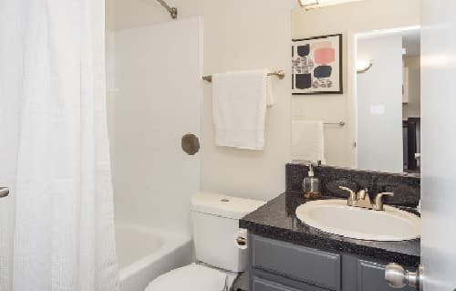 A small bathroom at The Social at Stadium Walk Fort Collins features a white shower curtain, white tiled shower, toilet, dark countertop with sink, towel on a rack, soap dispenser, and abstract wall art above the towel.